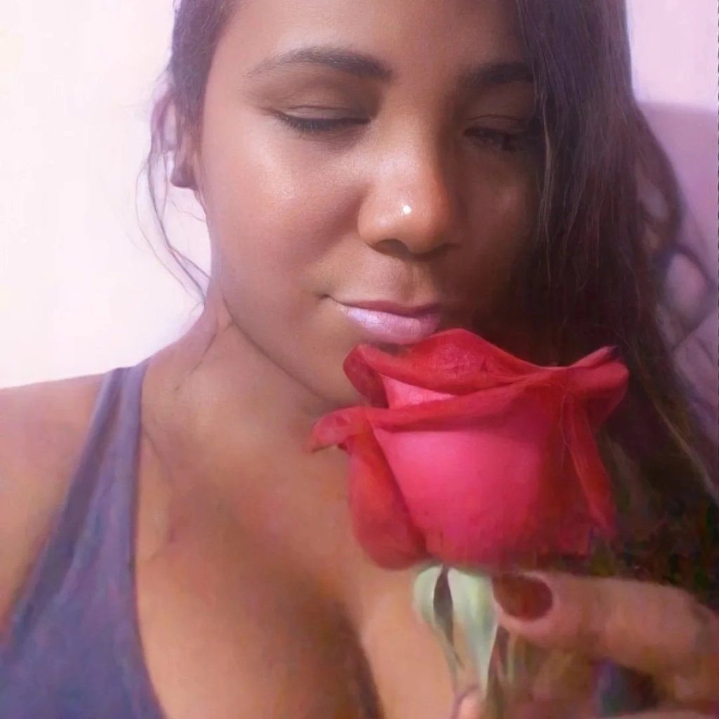 Uma foto em close de uma mulher com olhos fechados e expressão serena, segurando uma rosa vermelha aberta perto do rosto para cheirá-la. Ela tem pele morena e cabelos castanhos ondulados e usa uma blusa roxa, contra um fundo rosa suave e lilás.